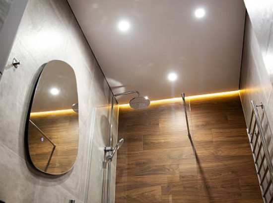Modern bathroom with wood tiles, mirror and stretch ceiling with LED lighting. Chrome shower and heated towel rail
