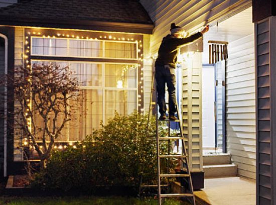 Woman on a ladder reaching up to a house soffit while hanging Christmas lights at night