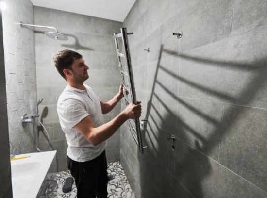 Side view of male plumber hanging electric towel rack dryer on the wall in bathroom. Man installing stainless steel bath towel heater in apartment under renovation. Home renovation concept.
